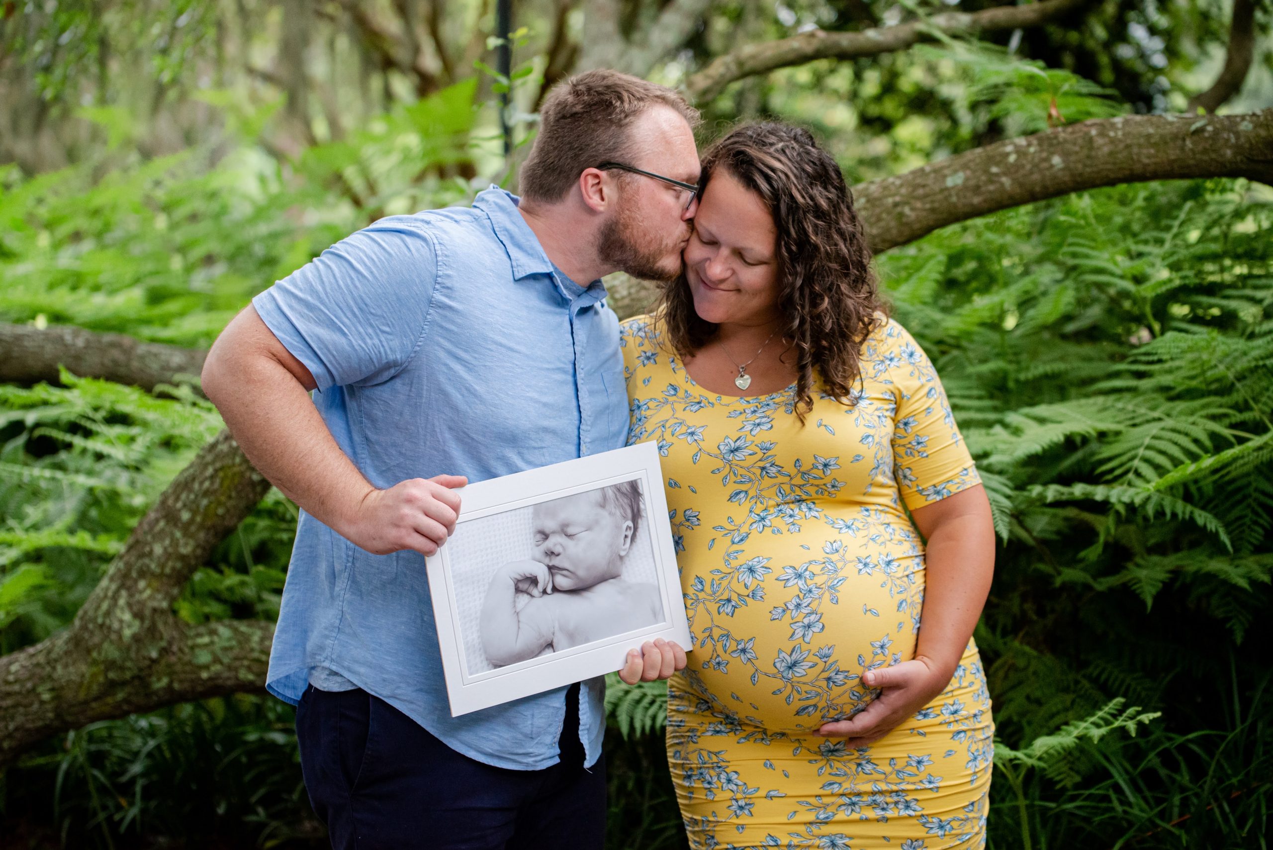 A dad-to-be kisses his pregnant wife on the cheek while holding an image of thier first baby who was born still