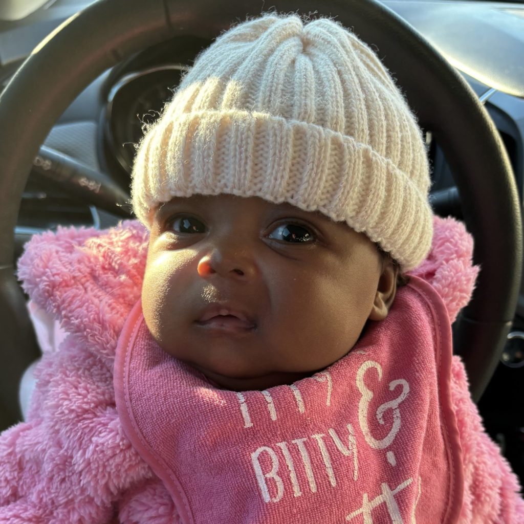 A baby in a pink fluffy outfit, wearing a cream knit hat and a bib