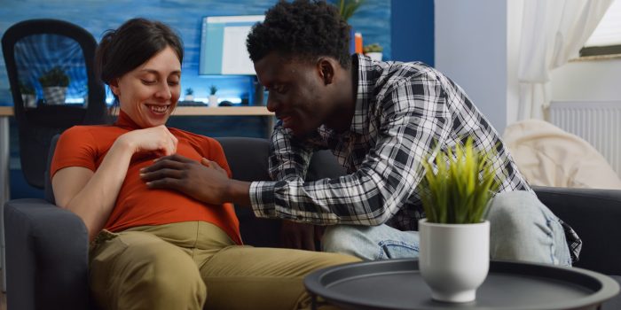 A young, interracial couple who is expecting a baby sit together on the sofa.