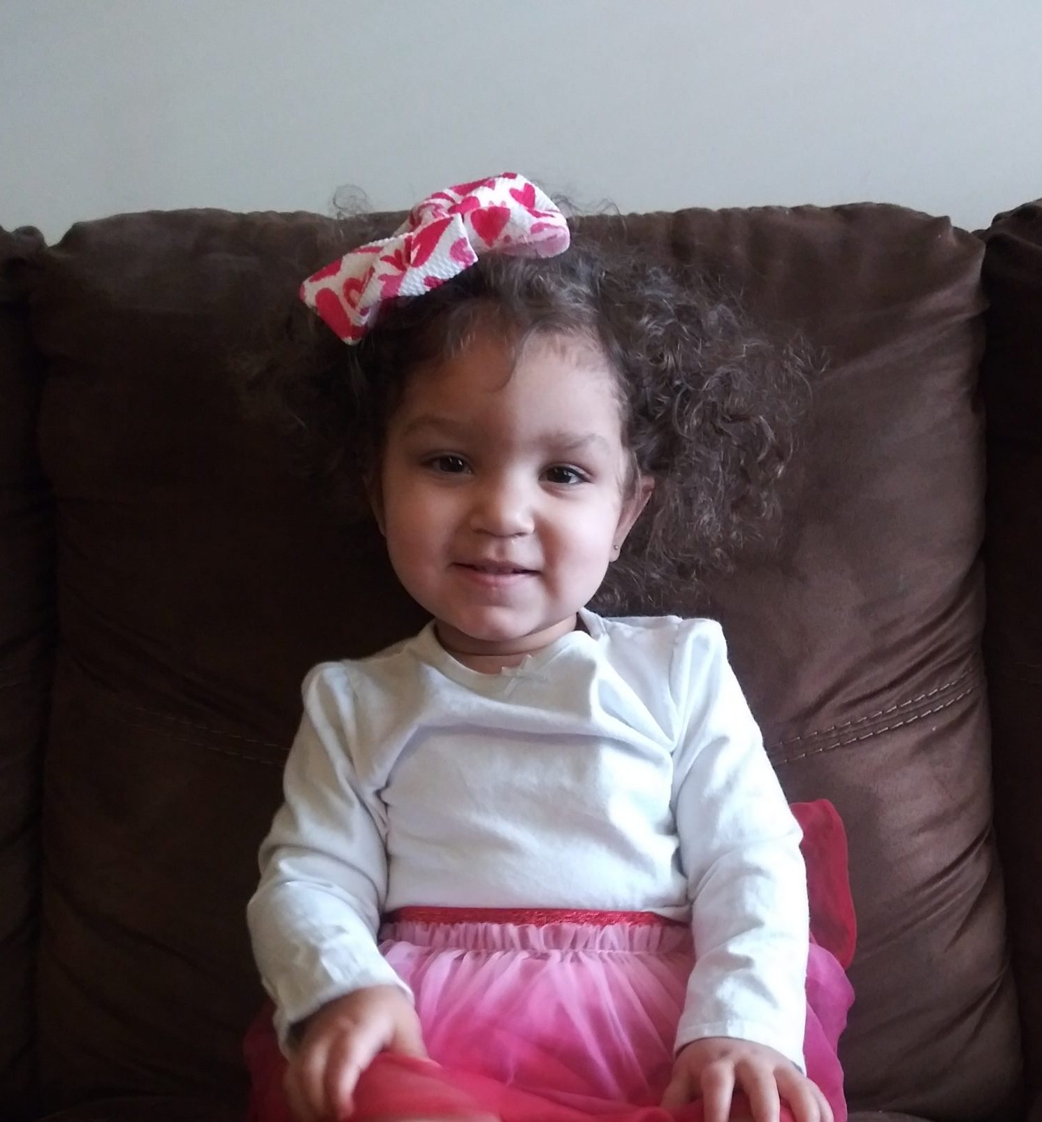 Baby save Nevaeh sits on a couch wearing a pink skirt and pink bow