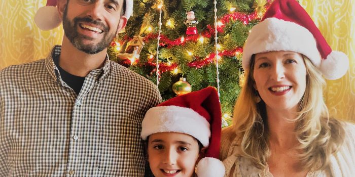 A family wearing Santa hats stand together in front of a Christmas tree