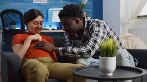 A young, interracial couple who is expecting a baby sit together on the sofa.