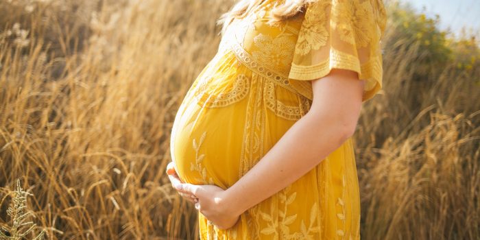 Pregnant woman in yellow dress standing in field resting her hands on her belly