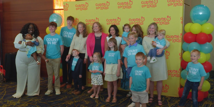 A group of adults and children, some wearing “Count on Me” shirts, pose in front of a “Count the Kicks” step-and-repeat banner and colorful balloons.