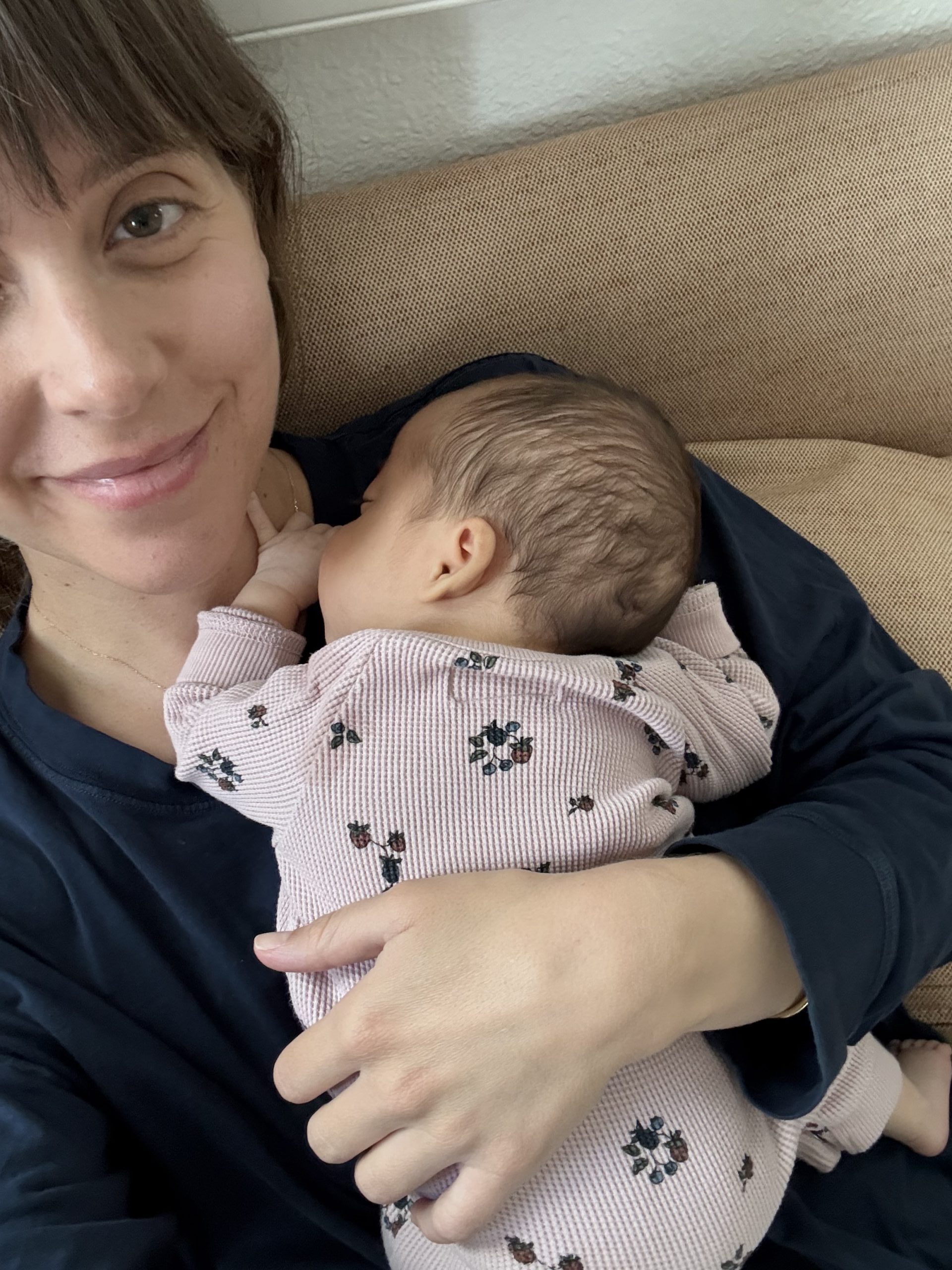 A woman smiles gently while holding a sleeping baby in her arms. The baby wears a floral-patterned outfit.