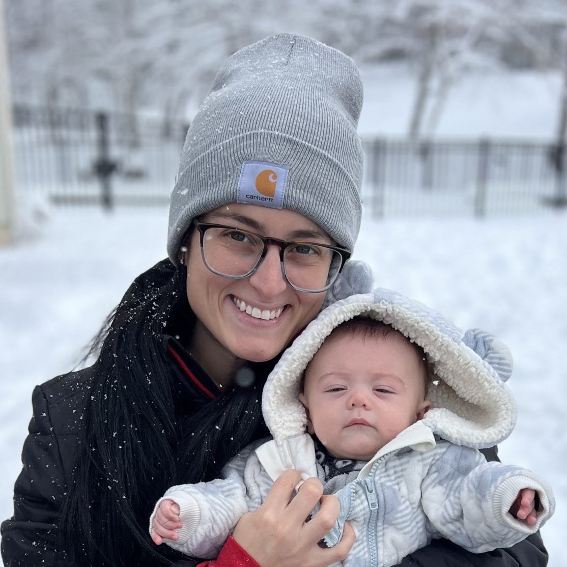A smiling woman in a gray beanie and black jacket holds a bundled baby in a snowy landscape.