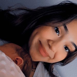 A woman smiles warmly at the camera while cradling a sleeping baby wrapped in a pink blanket.