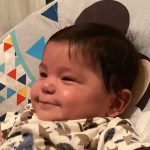 A baby wearing a patterned onesie smiles while lying in a colorful, geometric-patterned baby seat.