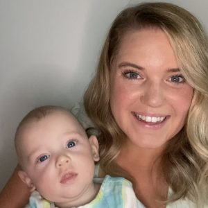 A smiling woman with long blonde hair holds a baby wearing a blue and yellow tie-dye outfit.