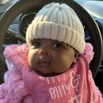 A baby in a pink fluffy outfit, wearing a cream knit hat and a bib