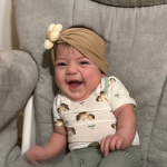 A smiling baby in a beige headband with a flower sits on a gray cushioned chair.