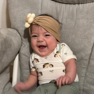 A smiling baby in a beige headband with a flower sits on a gray cushioned chair.