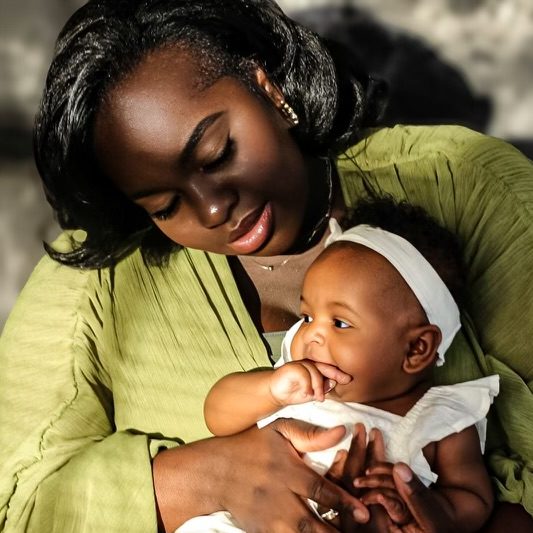 Woman in a green top lovingly holding a baby dressed in white with a headband.