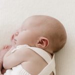 A newborn baby dressed in a white outfit lies curled up on a soft, white surface in a well-lit room.
