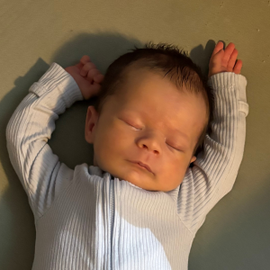 A newborn baby sleeps peacefully on a soft surface, wearing a light gray, ribbed onesie. The baby's arms are raised above their head.