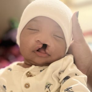 An infant wearing a white hat and shirt, sitting against a neutral background.