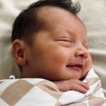 A sleeping newborn wrapped in a beige and white checkered blanket smiles gently. The baby rests peacefully on a soft, light-colored surface.