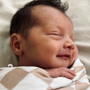 A sleeping newborn wrapped in a beige and white checkered blanket smiles gently. The baby rests peacefully on a soft, light-colored surface.