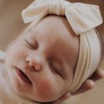 A baby wearing a cream-colored headband with a bow sleeps peacefully, resting against an adult hand for support.