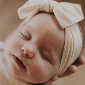 A baby wearing a cream-colored headband with a bow sleeps peacefully, resting against an adult hand for support.