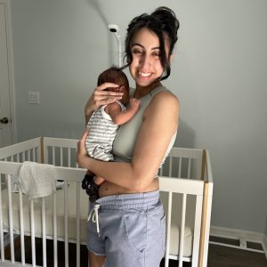A smiling woman holds a newborn baby in a nursery with a crib.