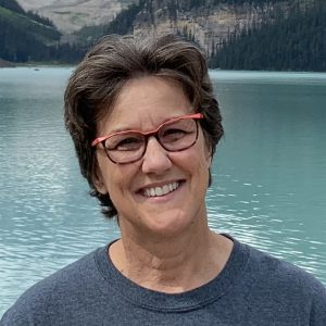 A smiling person with short brown hair and red-framed glasses wearing a dark gray shirt