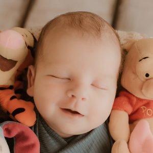 A sleeping newborn baby with a gentle smile, wrapped in a teal swaddle, lies in a cream-colored basket on a wooden floor.
