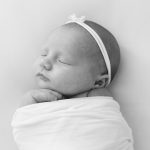 Black-and-white photo of a sleeping newborn baby wrapped in a white swaddle, wearing a white headband with a small bow.