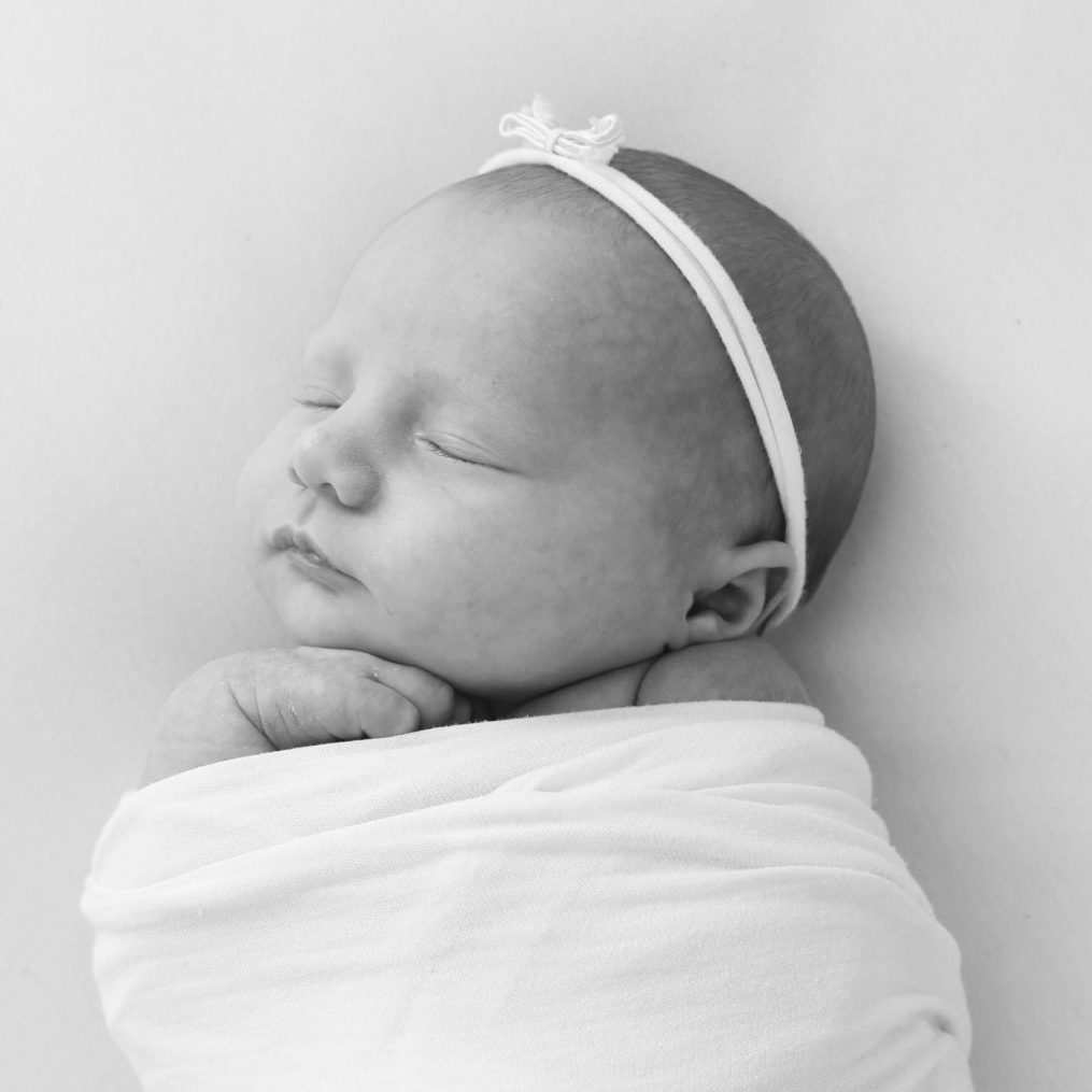 Black-and-white photo of a sleeping newborn baby wrapped in a white swaddle, wearing a white headband with a small bow.