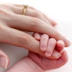 A close-up view of an adult hand with a gold ring gently holding the tiny, delicate hand of a newborn baby against a bright, soft white background.