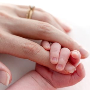 A close-up view of an adult hand with a gold ring gently holding the tiny, delicate hand of a newborn baby against a bright, soft white background.