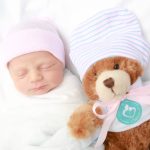 A newborn baby wrapped in a white blanket and wearing a pink hat sleeps beside a brown teddy bear dressed in a white outfit
